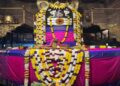 Close-up of the massive Shiva Lingam at Thanjavur Brihadeeswara Temple, decorated with colorful flower garlands, draped in pink and blue silk cloth, with a golden five-headed serpent (Naga) hood above it.