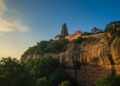 Majestic view of Tiruchengode Arthanareeswarar Temple situated on top of a red sandstone hill during sunset, surrounded by lush green trees.
