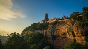 Majestic view of Tiruchengode Arthanareeswarar Temple situated on top of a red sandstone hill during sunset, surrounded by lush green trees.