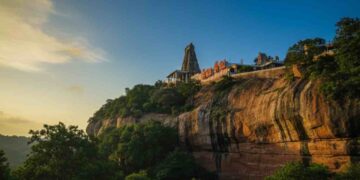Majestic view of Tiruchengode Arthanareeswarar Temple situated on top of a red sandstone hill during sunset, surrounded by lush green trees.