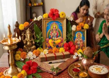 A detailed traditional Indian Hindu home altar prepared for Chitra Purnima, featuring a framed image of Lord Chitragupta, an idol of Lord Ganesha, and a framed picture of Lord Hanuman, all adorned with marigold and hibiscus flowers. The scene includes burning oil lamps, traditional ritual items on brass plates, and a bowl of sweet pudding, with blurred figures of people praying in the background.