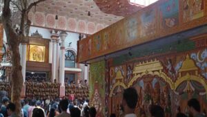 An interior shot of the Karya Siddhi Anjaneya Temple in Bangalore, showing a large crowd of devotees and ornate temple decorations.