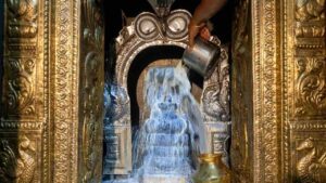 Close-up photograph of a milk abhishekam being performed on a Shiva Lingam at Thirupampuram Seshapureeswarar Temple, showing a hand pouring milk.