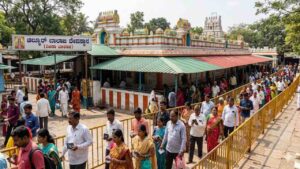A crowded dynamic outdoor scene at the historical Chilkur Balaji Temple, also known as the Visa Balaji Temple, in Hyderabad. Devotees are shown walking in an organized queue for pradakshina (circumambulation) along paved pathways, guided by low metal barriers. The iconic, colorful temple structure with its multiple towers, green and red corrugated roofs, and traditional yellow and blue finials is prominently featured under bright daylight, surrounded by trees. The main entrance features a clear sign with Kannada text 'ಚಿಲ್ಕೂರ್ ಬಾಲಾಜಿ ದೇವಸ್ಥಾನ (ವೀಸಾ ಬಾಲಾಜಿ)' (Chilkur Balaji Temple (Visa Balaji)). Several people are carrying small items like paper tokens, adding to the atmosphere of faith and devotion.