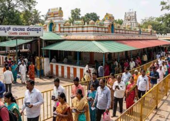 A crowded dynamic outdoor scene at the historical Chilkur Balaji Temple, also known as the Visa Balaji Temple, in Hyderabad. Devotees are shown walking in an organized queue for pradakshina (circumambulation) along paved pathways, guided by low metal barriers. The iconic, colorful temple structure with its multiple towers, green and red corrugated roofs, and traditional yellow and blue finials is prominently featured under bright daylight, surrounded by trees. The main entrance features a clear sign with Kannada text 'ಚಿಲ್ಕೂರ್ ಬಾಲಾಜಿ ದೇವಸ್ಥಾನ (ವೀಸಾ ಬಾಲಾಜಿ)' (Chilkur Balaji Temple (Visa Balaji)). Several people are carrying small items like paper tokens, adding to the atmosphere of faith and devotion.