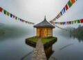 A wide-angle landscape shot of a small wooden temple with a pagoda-style roof sitting on a tiny island in a serene, misty mountain lake. Stone pathways lined with wire mesh lead to it, and colorful Tibetan prayer flags hang overhead against the cloudy sky. Green hills and forests form the background, reflected in the still water.