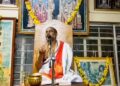 Sri Brahmanyachar delivering a spiritual lecture on Adhika Masa importance in Mantralaya, seated in front of deities' photos.