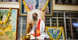Sri Brahmanyachar delivering a spiritual lecture on Adhika Masa importance in Mantralaya, seated in front of deities' photos.