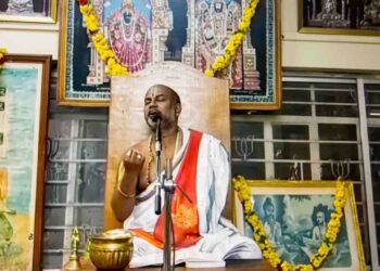 Sri Brahmanyachar delivering a spiritual lecture on Adhika Masa importance in Mantralaya, seated in front of deities' photos.
