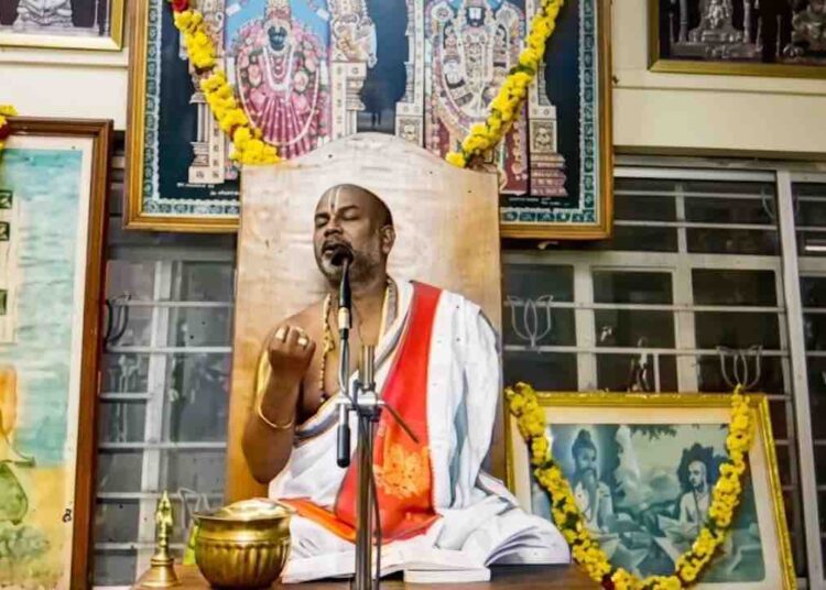 Sri Brahmanyachar delivering a spiritual lecture on Adhika Masa importance in Mantralaya, seated in front of deities' photos.