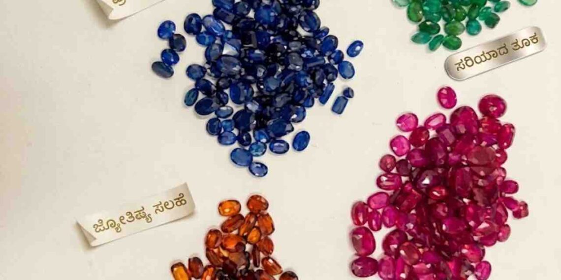 Top-down photograph showing four neat piles of faceted oval-cut gemstones arranged diagonally on an off-white surface. The clusters, which are in hues of sapphire blue, emerald green, ruby red, and spessartite orange, are each paired with a delicate Kannada-text gold label. In the bottom-right corner, the JS Gems & Jewellery logo and certification text in English and Kannada are displayed.