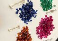 Top-down photograph showing four neat piles of faceted oval-cut gemstones arranged diagonally on an off-white surface. The clusters, which are in hues of sapphire blue, emerald green, ruby red, and spessartite orange, are each paired with a delicate Kannada-text gold label. In the bottom-right corner, the JS Gems & Jewellery logo and certification text in English and Kannada are displayed.