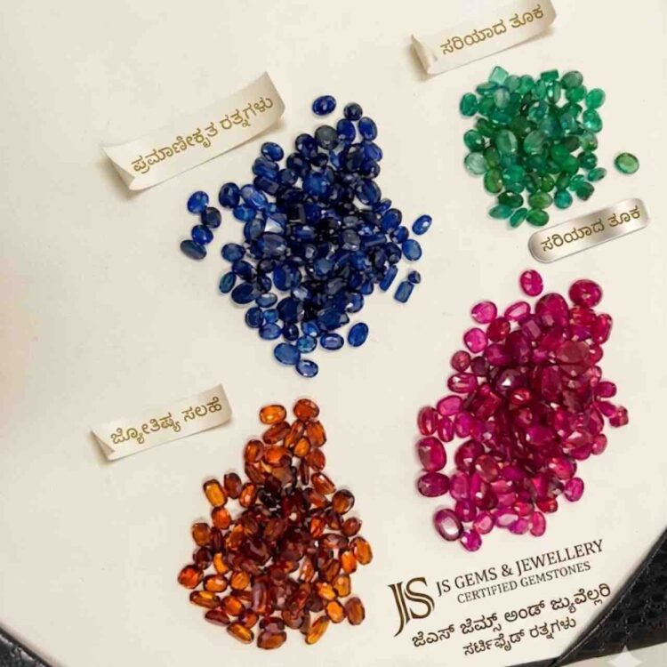 Top-down photograph showing four neat piles of faceted oval-cut gemstones arranged diagonally on an off-white surface. The clusters, which are in hues of sapphire blue, emerald green, ruby red, and spessartite orange, are each paired with a delicate Kannada-text gold label. In the bottom-right corner, the JS Gems & Jewellery logo and certification text in English and Kannada are displayed.