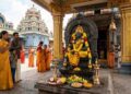 A divine South Indian temple scene where devotees in yellow traditional attire are praying to Lord Guru (Brihaspati) idol decorated with yellow flowers.