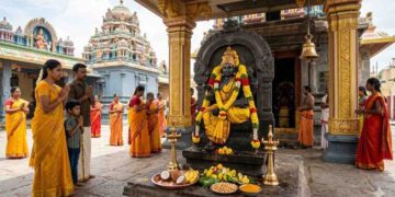 A divine South Indian temple scene where devotees in yellow traditional attire are praying to Lord Guru (Brihaspati) idol decorated with yellow flowers.