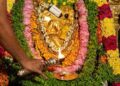 Close-up photograph of a golden idol of Lord Bhuvaraha, the boar-headed incarnation of Vishnu, at Tirumala Tirupati Bhuvaraha Temple. The idol is richly decorated with flower garlands. A priest performs arati with a multi-wick lamp, and a devotee offers prayers with a spoon, as traditional lamps burn in the background.