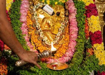 Close-up photograph of a golden idol of Lord Bhuvaraha, the boar-headed incarnation of Vishnu, at Tirumala Tirupati Bhuvaraha Temple. The idol is richly decorated with flower garlands. A priest performs arati with a multi-wick lamp, and a devotee offers prayers with a spoon, as traditional lamps burn in the background.