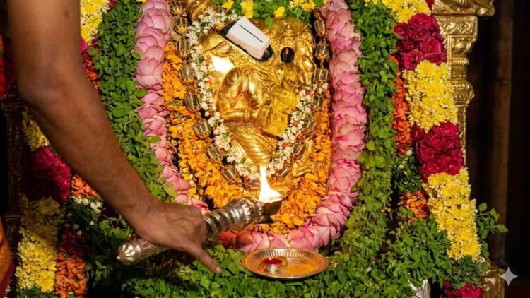 Close-up photograph of a golden idol of Lord Bhuvaraha, the boar-headed incarnation of Vishnu, at Tirumala Tirupati Bhuvaraha Temple. The idol is richly decorated with flower garlands. A priest performs arati with a multi-wick lamp, and a devotee offers prayers with a spoon, as traditional lamps burn in the background.