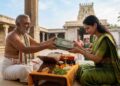 A woman in a traditional green saree donating the Shrimad Bhagavata Mahapurana book to a traditional Hindu scholar in a temple courtyard.