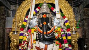 Close-up HD shot of the historic Kurudumale Ganesha idol decorated with vibrant flower garlands and a golden arch in the background.