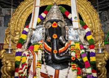 Close-up HD shot of the historic Kurudumale Ganesha idol decorated with vibrant flower garlands and a golden arch in the background.