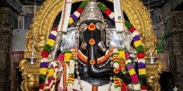 Close-up HD shot of the historic Kurudumale Ganesha idol decorated with vibrant flower garlands and a golden arch in the background.