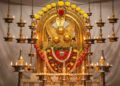 A high-definition photograph of the main deity (Thidambu) of the Taliparamba Rajarajeswara Temple, intricately decorated with gold ornaments, yellow roses, and red garlands. The deity is surrounded by numerous traditional oil lamps and set against a white background.