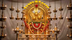 A high-definition photograph of the main deity (Thidambu) of the Taliparamba Rajarajeswara Temple, intricately decorated with gold ornaments, yellow roses, and red garlands. The deity is surrounded by numerous traditional oil lamps and set against a white background.