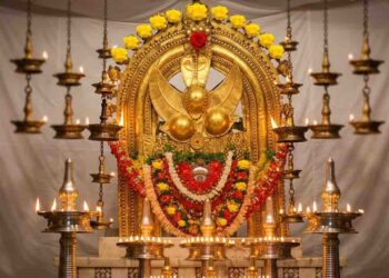 A high-definition photograph of the main deity (Thidambu) of the Taliparamba Rajarajeswara Temple, intricately decorated with gold ornaments, yellow roses, and red garlands. The deity is surrounded by numerous traditional oil lamps and set against a white background.