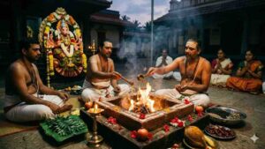 A realistic image of a Varahi Homam (sacrificial fire ritual) being performed. Priests are seated around a square fire pit (Homa Kunda), offering materials into the flames. In the background, there is a decorated idol of Goddess Varahi (with a boar face) and devotees seated. Various ritual items like coconuts, pomegranates, and dried red chilies are visible in the foreground.