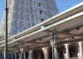 A low-angle shot of the towering, white, ornate gopuram (temple tower) of the Tiruchendur Murugan Temple, viewed from a paved courtyard with devotees in the foreground.