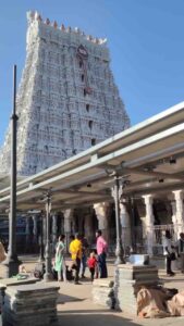 A low-angle shot of the towering, white, ornate gopuram (temple tower) of the Tiruchendur Murugan Temple, viewed from a paved courtyard with devotees in the foreground.