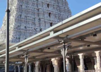 A low-angle shot of the towering, white, ornate gopuram (temple tower) of the Tiruchendur Murugan Temple, viewed from a paved courtyard with devotees in the foreground.