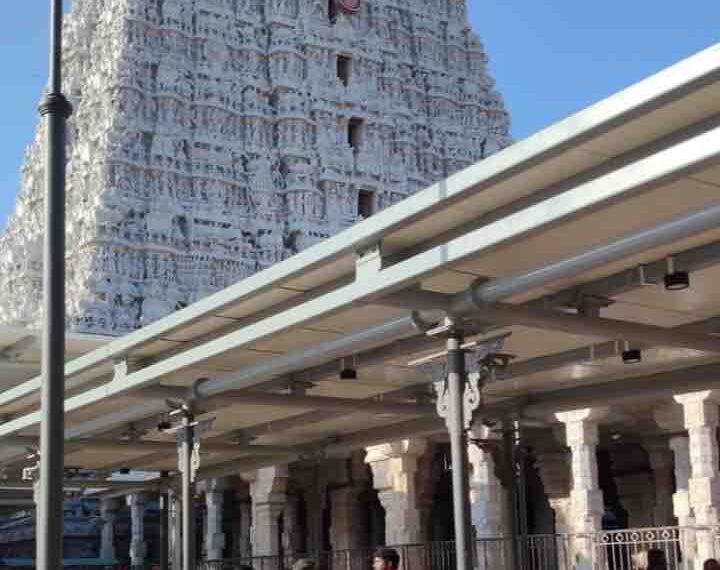 A low-angle shot of the towering, white, ornate gopuram (temple tower) of the Tiruchendur Murugan Temple, viewed from a paved courtyard with devotees in the foreground.