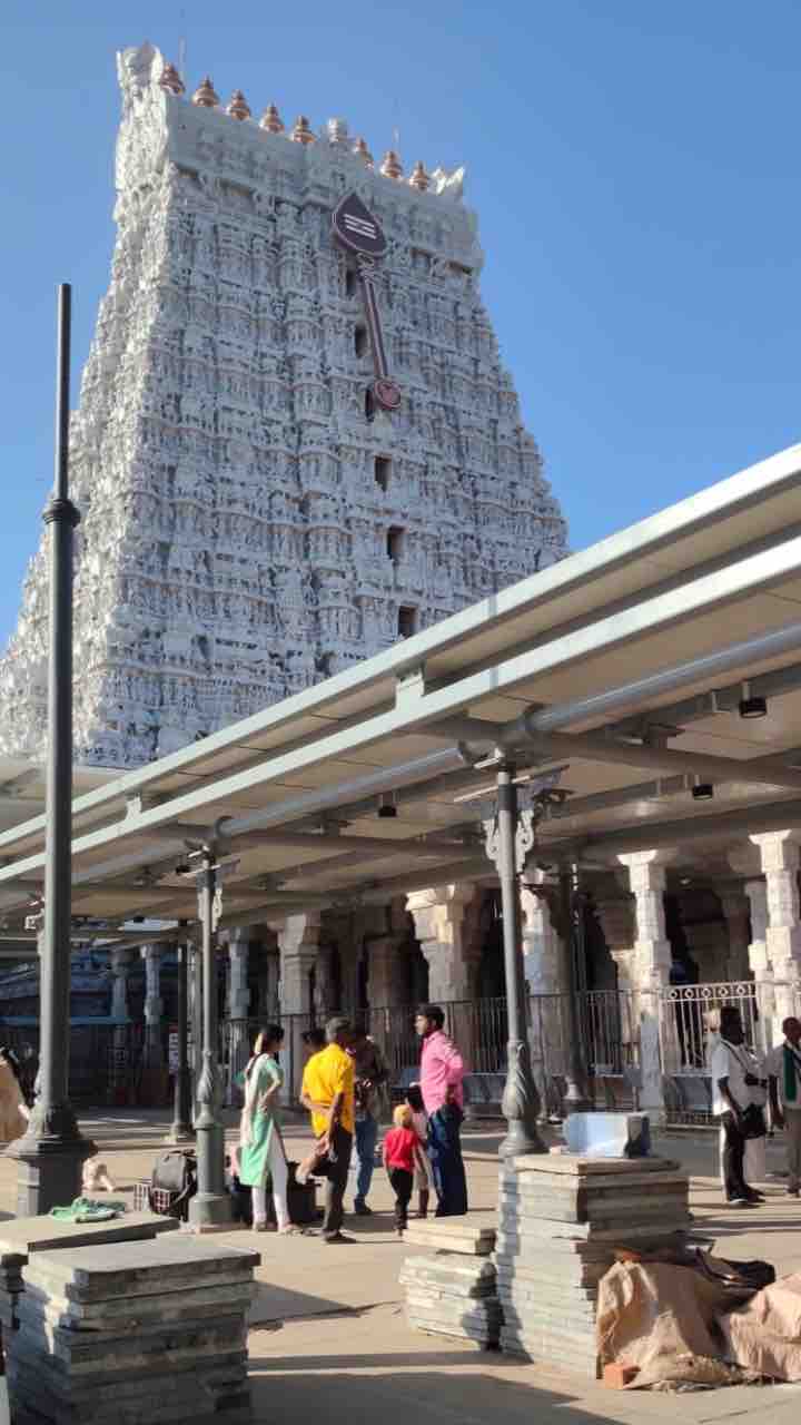 A low-angle shot of the towering, white, ornate gopuram (temple tower) of the Tiruchendur Murugan Temple, viewed from a paved courtyard with devotees in the foreground.