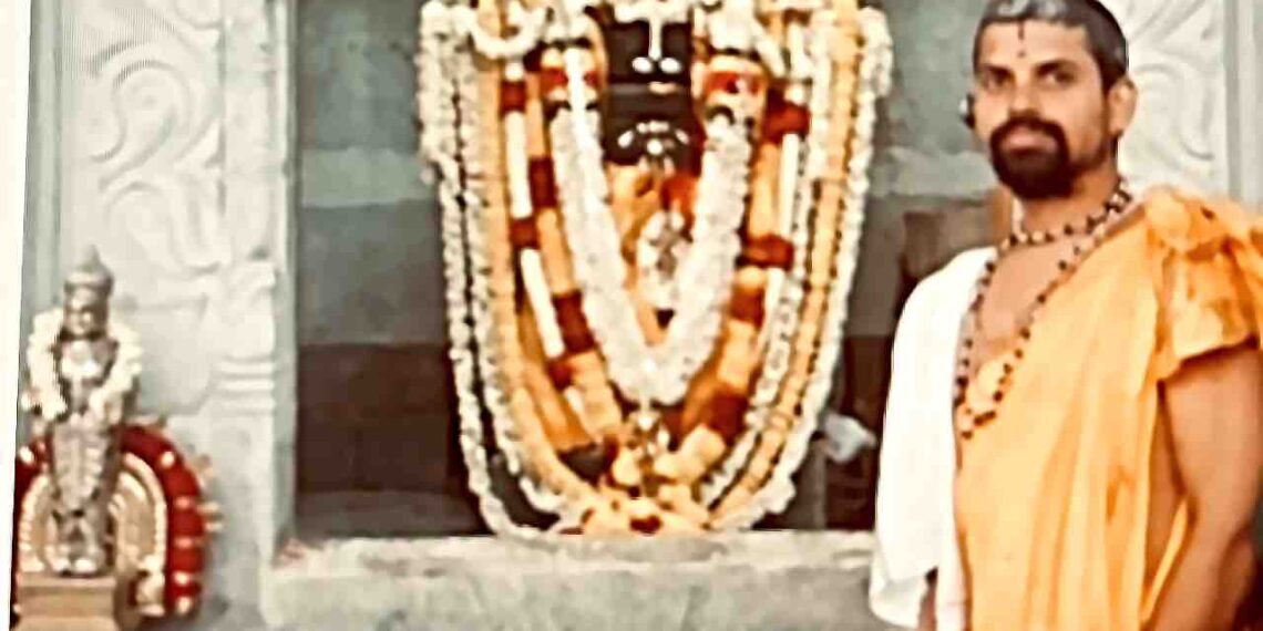 A priest in traditional saffron attire and rudraksha beads standing beside a garlanded deity shrine (vrindavana) at a temple, with intricately carved stone pillars in the background.