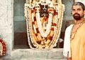 A priest in traditional saffron attire and rudraksha beads standing beside a garlanded deity shrine (vrindavana) at a temple, with intricately carved stone pillars in the background.