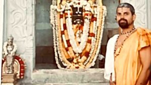 A priest in traditional saffron attire and rudraksha beads standing beside a garlanded deity shrine (vrindavana) at a temple, with intricately carved stone pillars in the background.