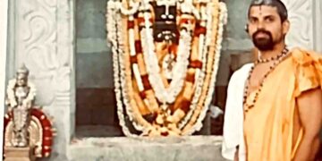 A priest in traditional saffron attire and rudraksha beads standing beside a garlanded deity shrine (vrindavana) at a temple, with intricately carved stone pillars in the background.