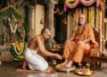 A disciple kneels and performs Pada Puja, washing the feet of a seated Hindu Guru with flowers and water during Guru Purnima.