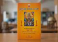 ⁠A close-up photograph of the vibrant orange cover of a Kannada astrology book titled 'Jyotishya Udaya Deepika' by K. Prakash Rao. The cover features a central illustration of Lord Surya in a seven-horse chariot, flanked by oil lamps, with details of the author's title and the publisher at the bottom. The book stands vertically on a wooden desk with a blurred library background.