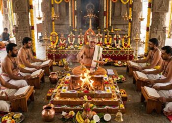 An authentic Vedic ritual in an ornate South Indian temple where a group of barefoot Hindu priests, clad in traditional white and gold clothing with bare torsos and distinct forehead markings, are performing a Navagraha Homa around a central sacred fire (Homa Kunda).