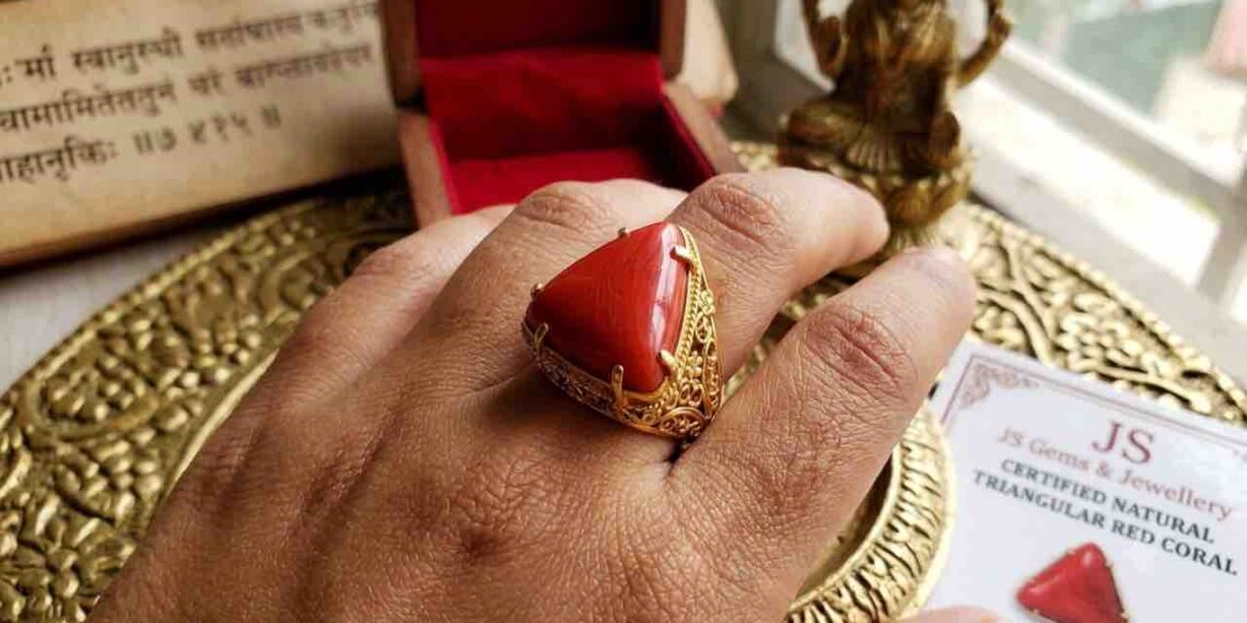 A close-up photograph of a left hand wearing a triangular red coral (Lakshmi Havala) gemstone set in an intricate gold filigree ring on the ring finger.