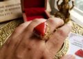 A close-up photograph of a left hand wearing a triangular red coral (Lakshmi Havala) gemstone set in an intricate gold filigree ring on the ring finger.