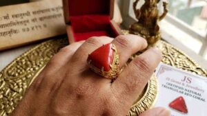A close-up photograph of a left hand wearing a triangular red coral (Lakshmi Havala) gemstone set in an intricate gold filigree ring on the ring finger.