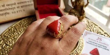 A close-up photograph of a left hand wearing a triangular red coral (Lakshmi Havala) gemstone set in an intricate gold filigree ring on the ring finger.