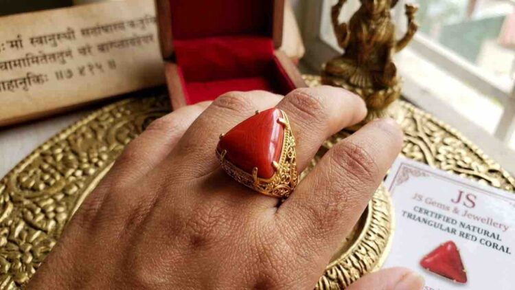 A close-up photograph of a left hand wearing a triangular red coral (Lakshmi Havala) gemstone set in an intricate gold filigree ring on the ring finger.