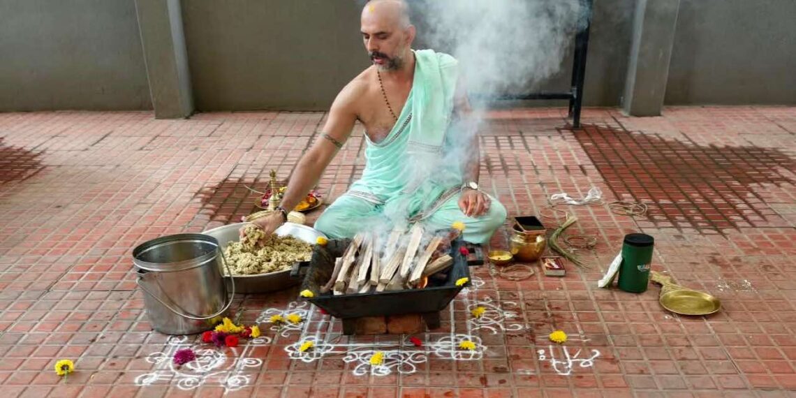 A photo showing Purohit Manjunath N Bharadwaj performing the Akhanda Maha Ganapathi Homa at the Shirdi Sai Baba Mandir premises in Bangalore. He is seated on the floor, surrounded by ceremonial items like a metal bucket, flowers, and a brass pot, while placing offerings into a square fire pit from which smoke is rising. He is wearing a light mint-green traditional attire and has a watch on his left wrist.