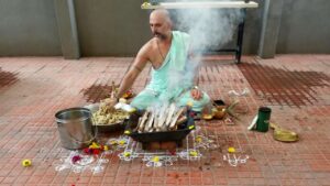 A photo showing Purohit Manjunath N Bharadwaj performing the Akhanda Maha Ganapathi Homa at the Shirdi Sai Baba Mandir premises in Bangalore. He is seated on the floor, surrounded by ceremonial items like a metal bucket, flowers, and a brass pot, while placing offerings into a square fire pit from which smoke is rising. He is wearing a light mint-green traditional attire and has a watch on his left wrist.
