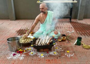 A photo showing Purohit Manjunath N Bharadwaj performing the Akhanda Maha Ganapathi Homa at the Shirdi Sai Baba Mandir premises in Bangalore. He is seated on the floor, surrounded by ceremonial items like a metal bucket, flowers, and a brass pot, while placing offerings into a square fire pit from which smoke is rising. He is wearing a light mint-green traditional attire and has a watch on his left wrist.